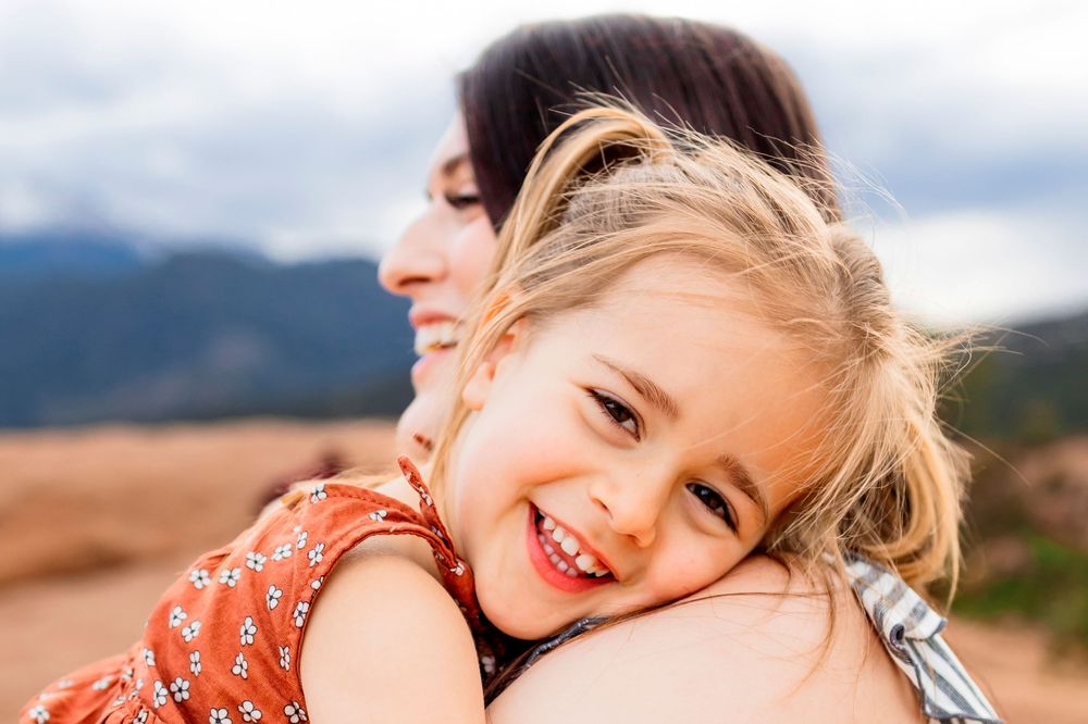 A toddler girl placing her head on her mothers shoulder.  She is looking at the camera and smiling