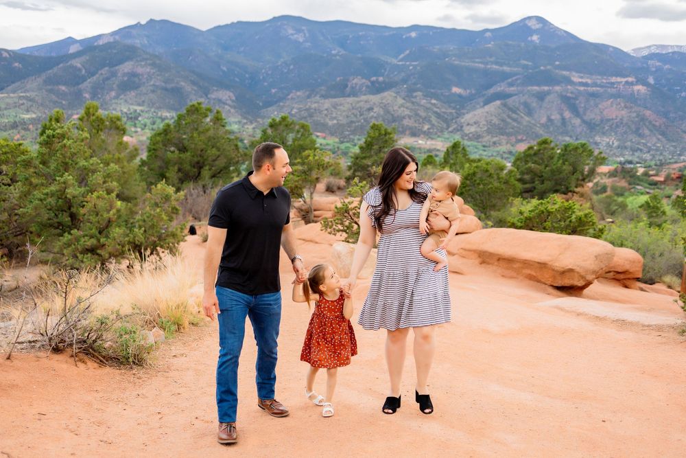 A family of four walks along a scenic red rock trail overlooking mountains and valleys in Garden of the Gods, Colorado.