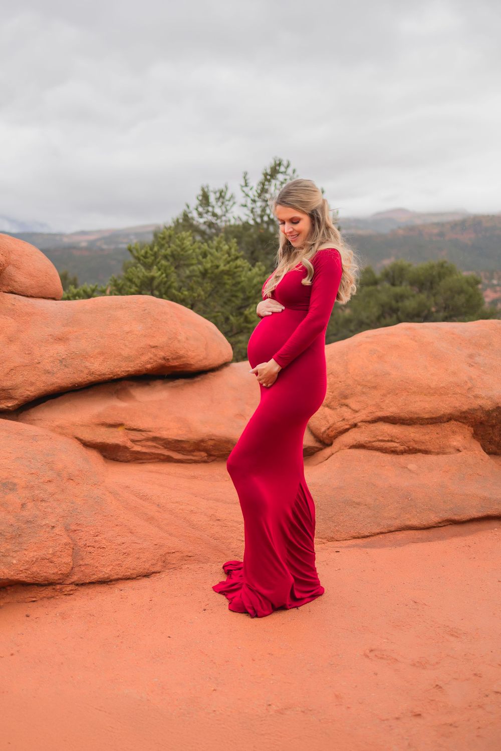 A pregnant woman in a flowing red dress poses gracefully against the striking orange sandstone formations at Garden of the Gods.