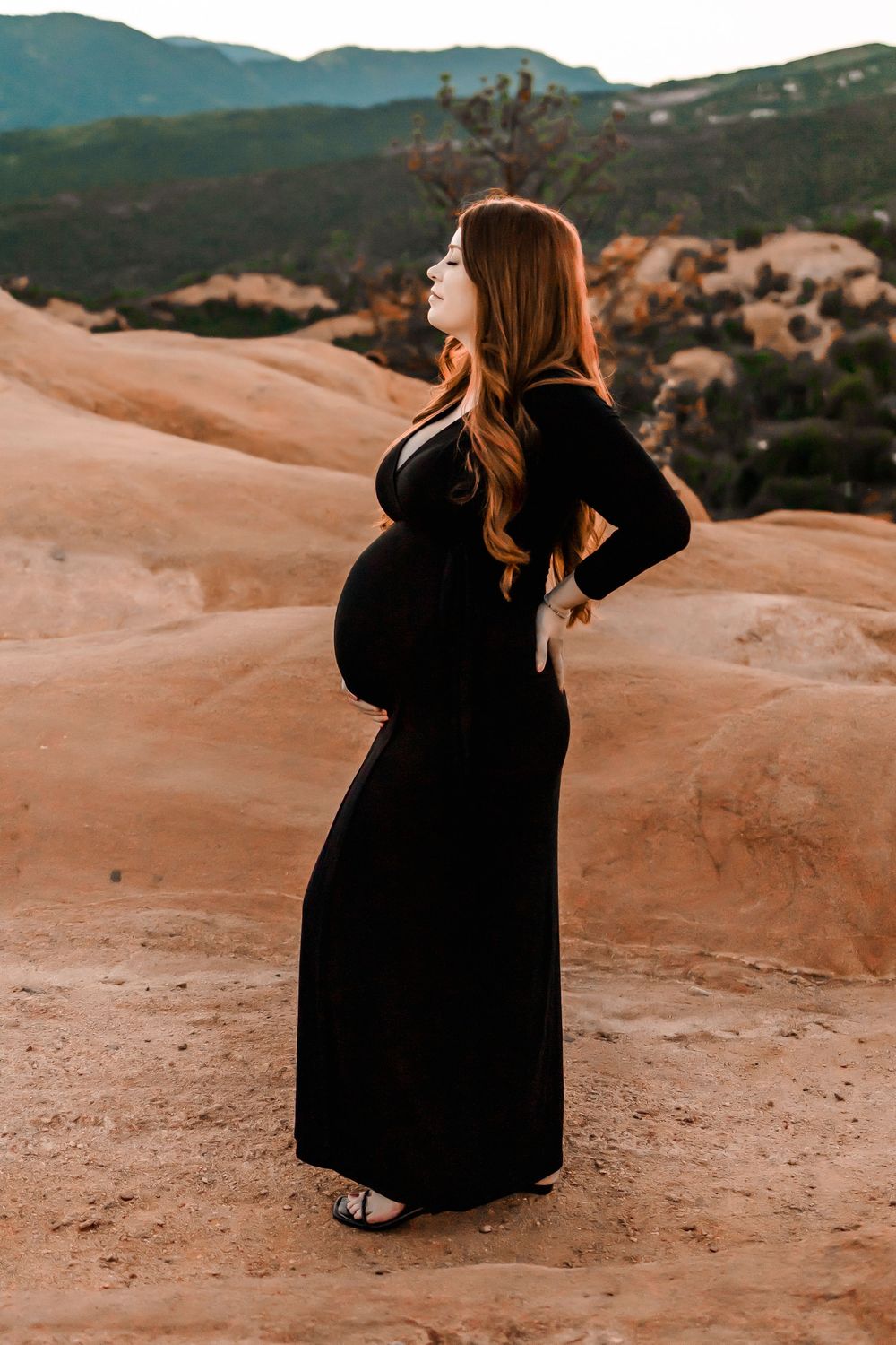 A pregnant woman in a flowing black dress poses  against orange clay hills and mountains of garden of the gods at sunset.