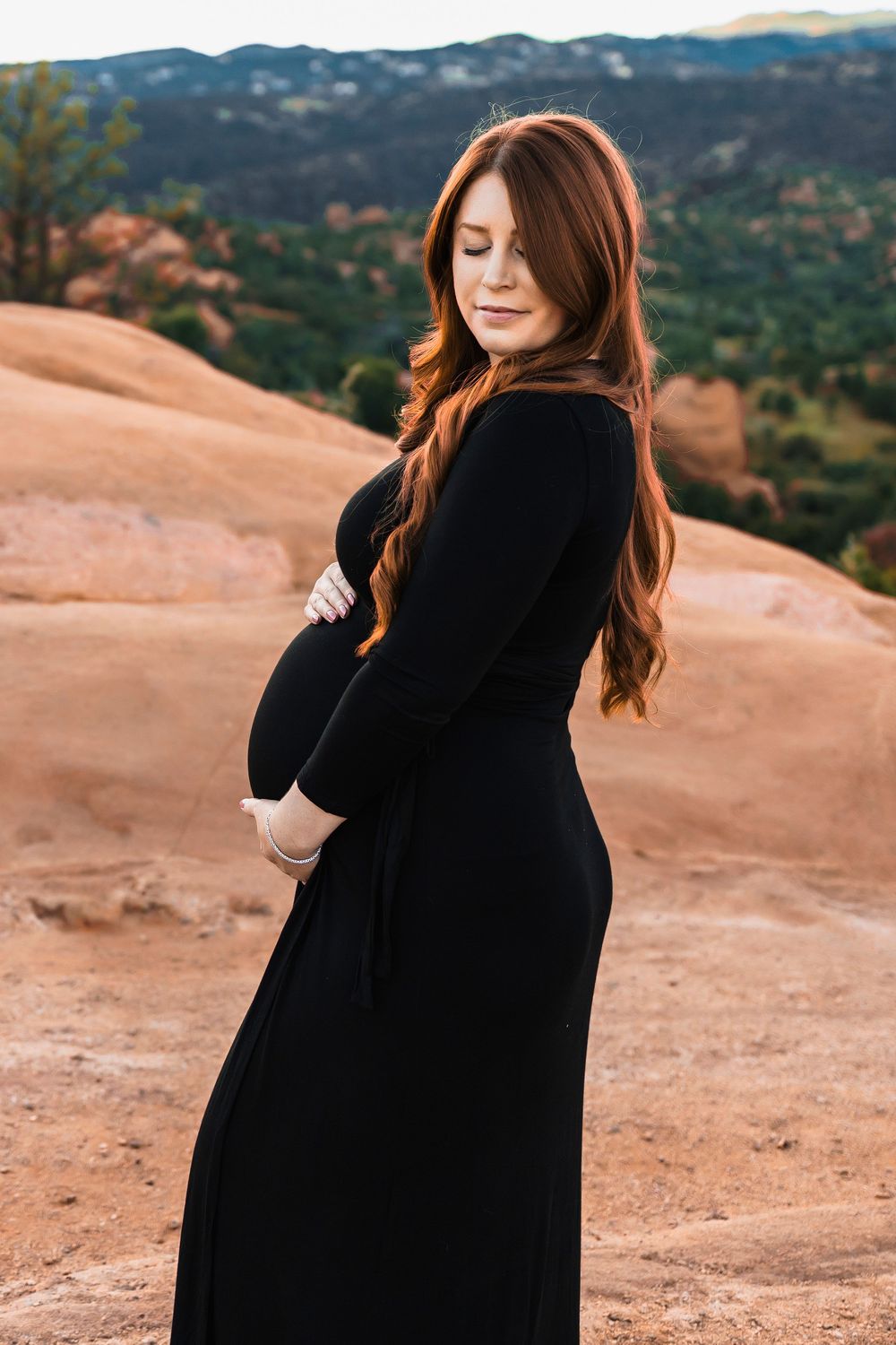 A pregnant woman in a long black dress poses against red rocks and mountain scenery at sunset at garden of the gods