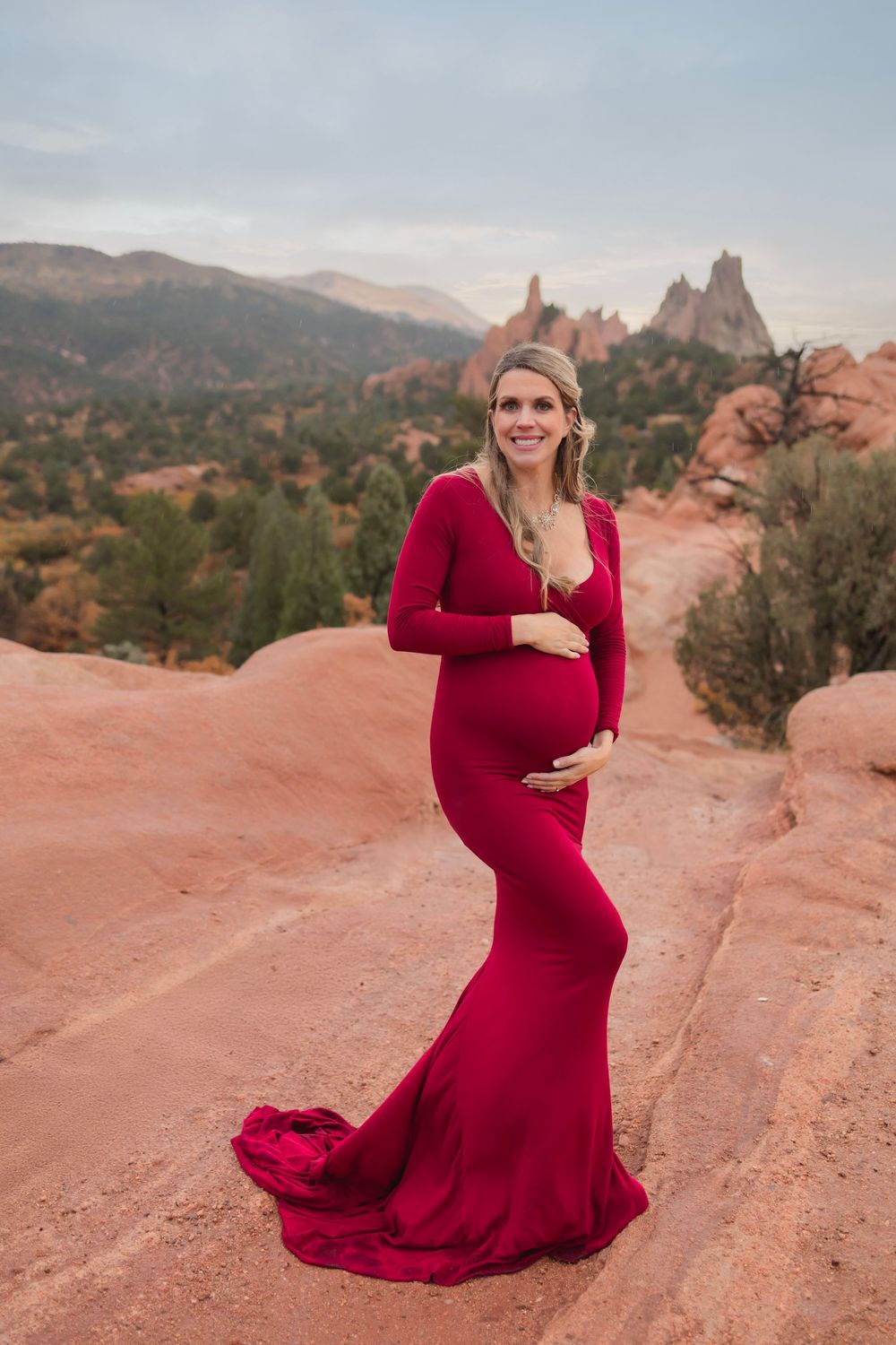 A pregnant woman in a flowing red maternity gown poses on red rocks at Garden of the Gods during sunset.
