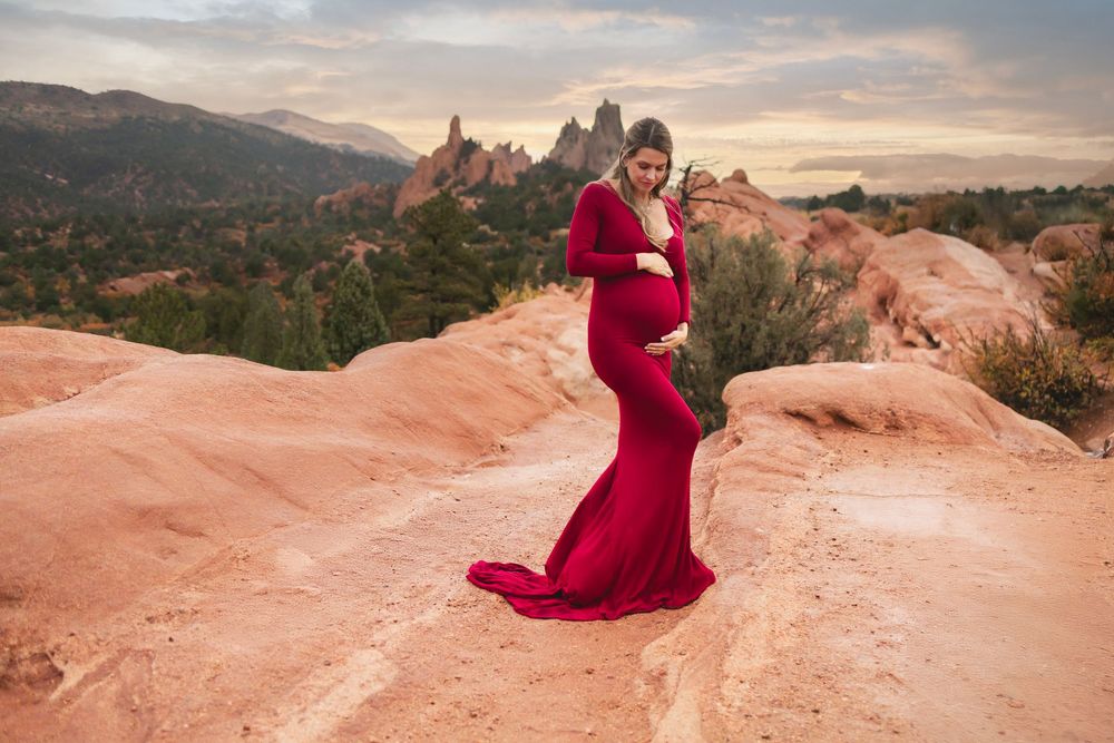 A pregnant woman in flowing red dress contrasts against orange sandstone formations at Garden of the Gods during sunset.
