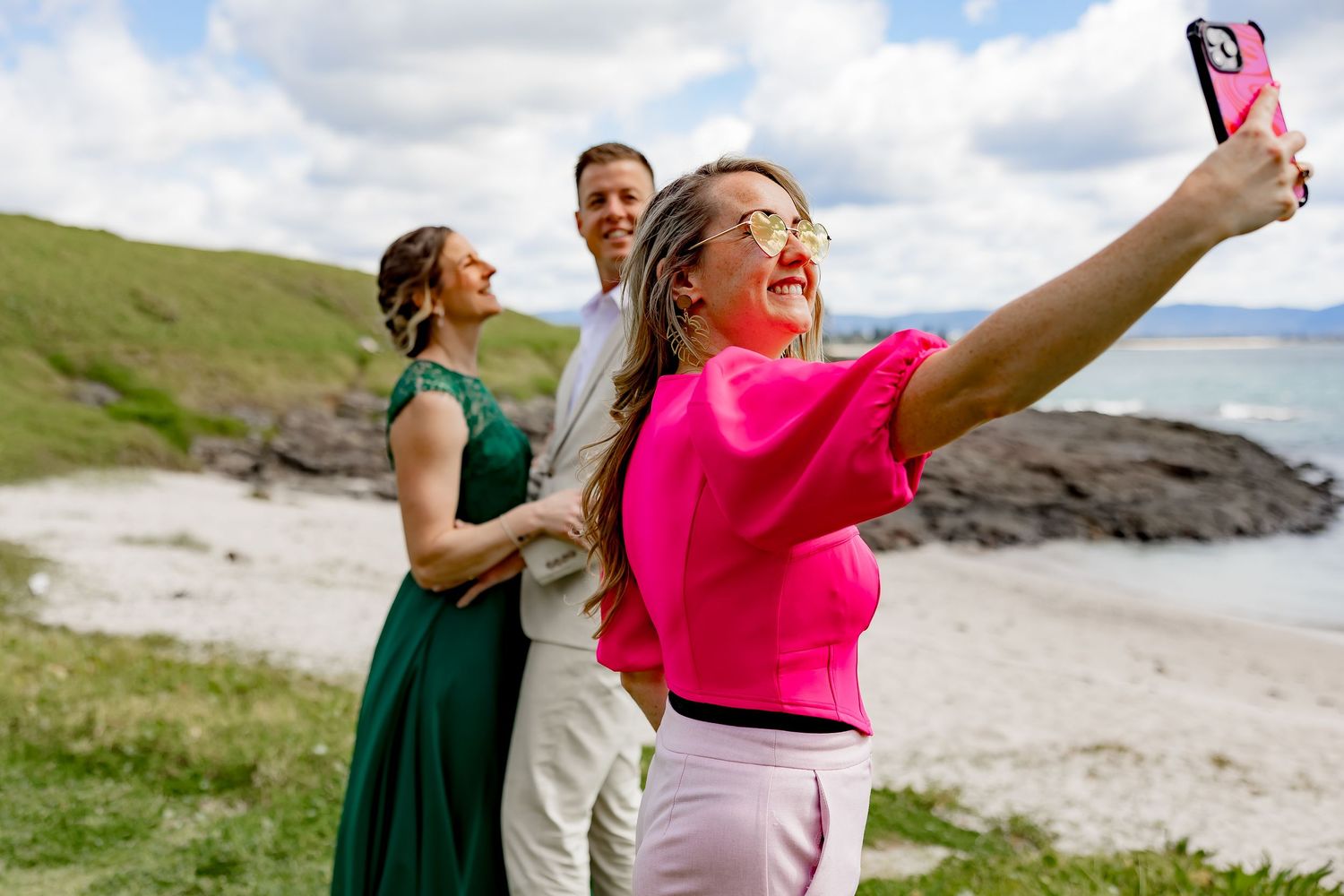 Friends enjoying a fun moment taking selfies on a scenic beach with green hills and rocky coastline in the background.