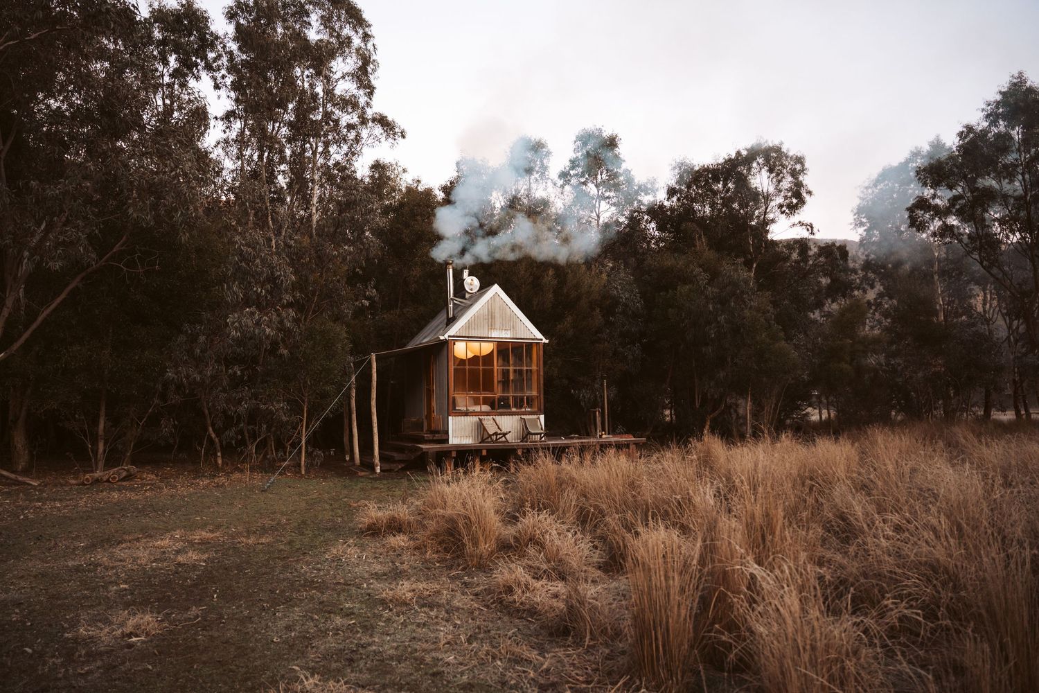 Small wooden cabin with illuminated windows and chimney smoke at dusk, surrounded by golden grassland and eucalyptus forest in rural Austral