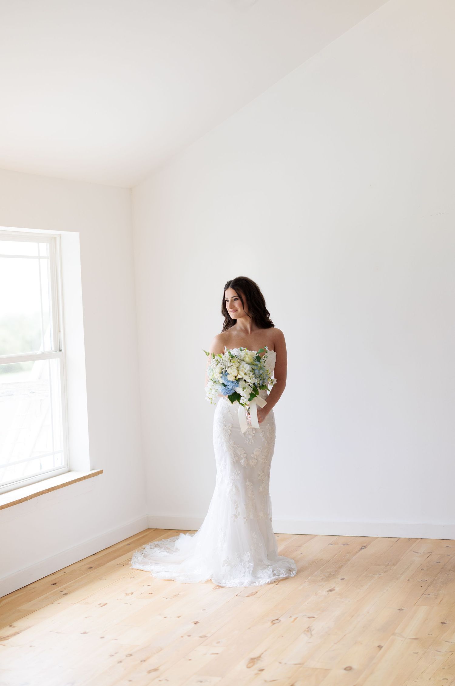 Bride in white lace gown holds blue and white bouquet in minimalist room with window.