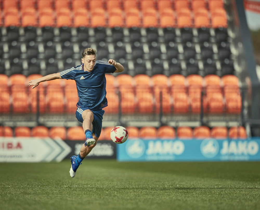 Soccer player kicking ball on field with stadium seats in background.