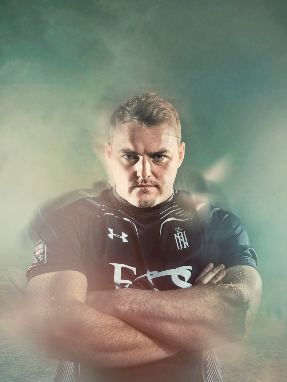 Athlete in dark sports uniform poses with crossed arms in dramatic lighting.