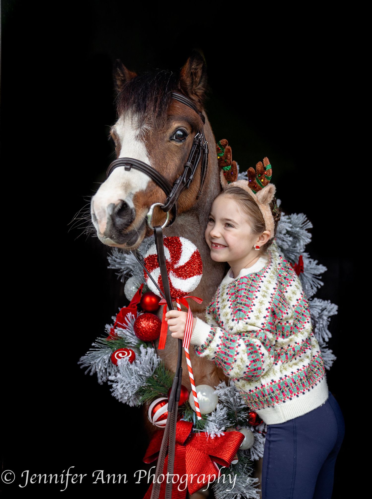 A festive holiday photo shoot features a pony with a red bow and silver tinsel garland against a black background.