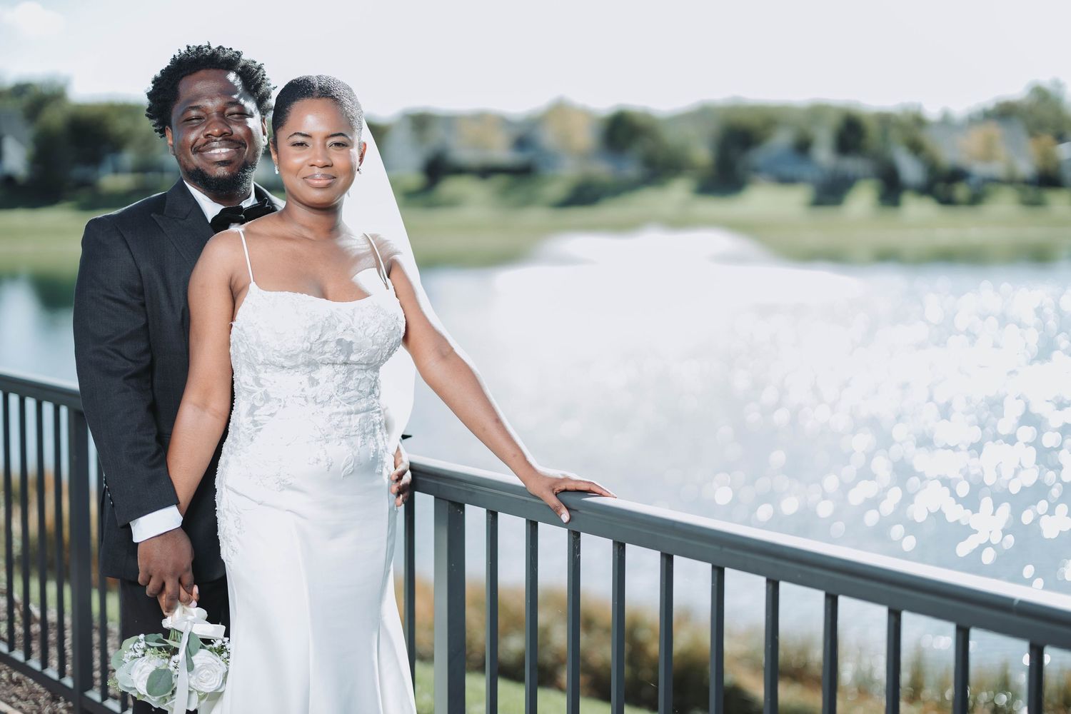 A couple in wedding attire poses together on a waterfront deck during a sunny outdoor celebration.