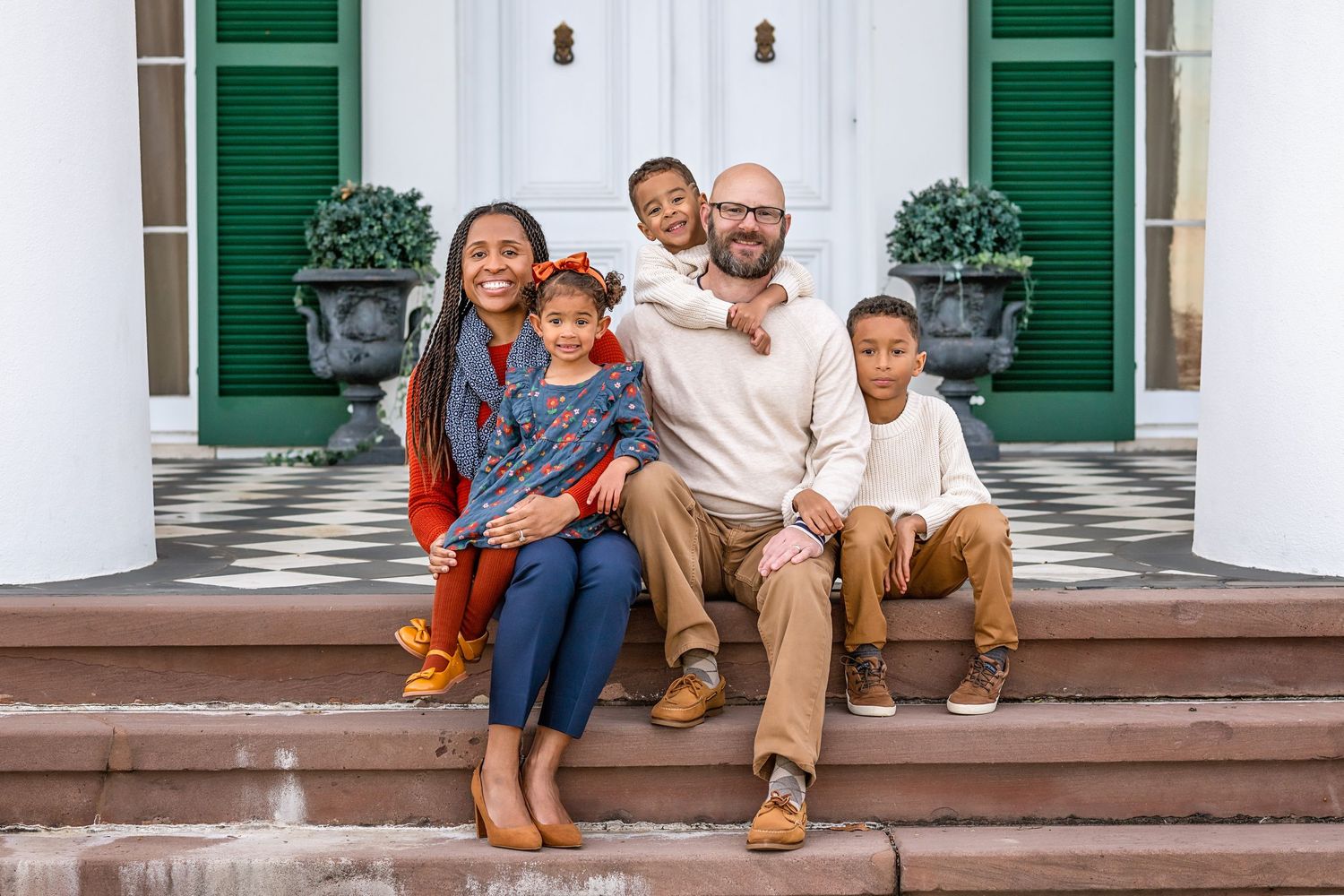 Family wearing coordinated fall outfits sits together on white porch steps with green shutters in background.