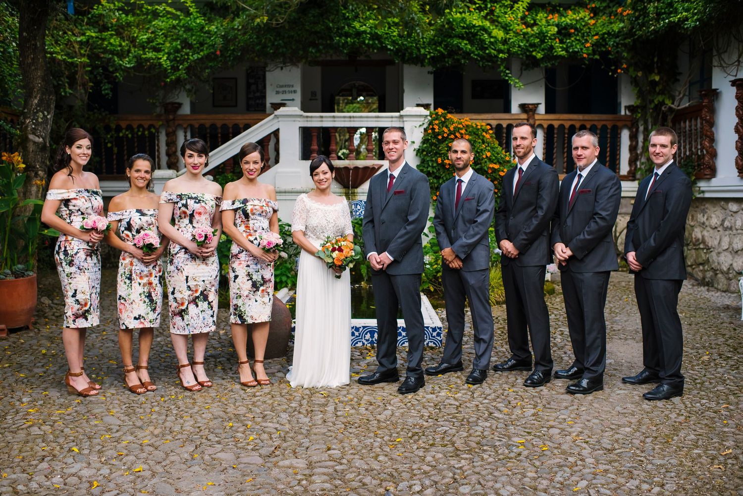 Wedding party poses outdoors with bridesmaids in floral dresses and groomsmen in dark suits.