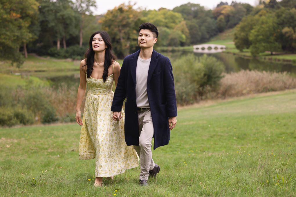 An engagement shoot of a couple walking together through a scenic grassy meadow on a warm autumn afternoon. Painshill Park in Surrey.