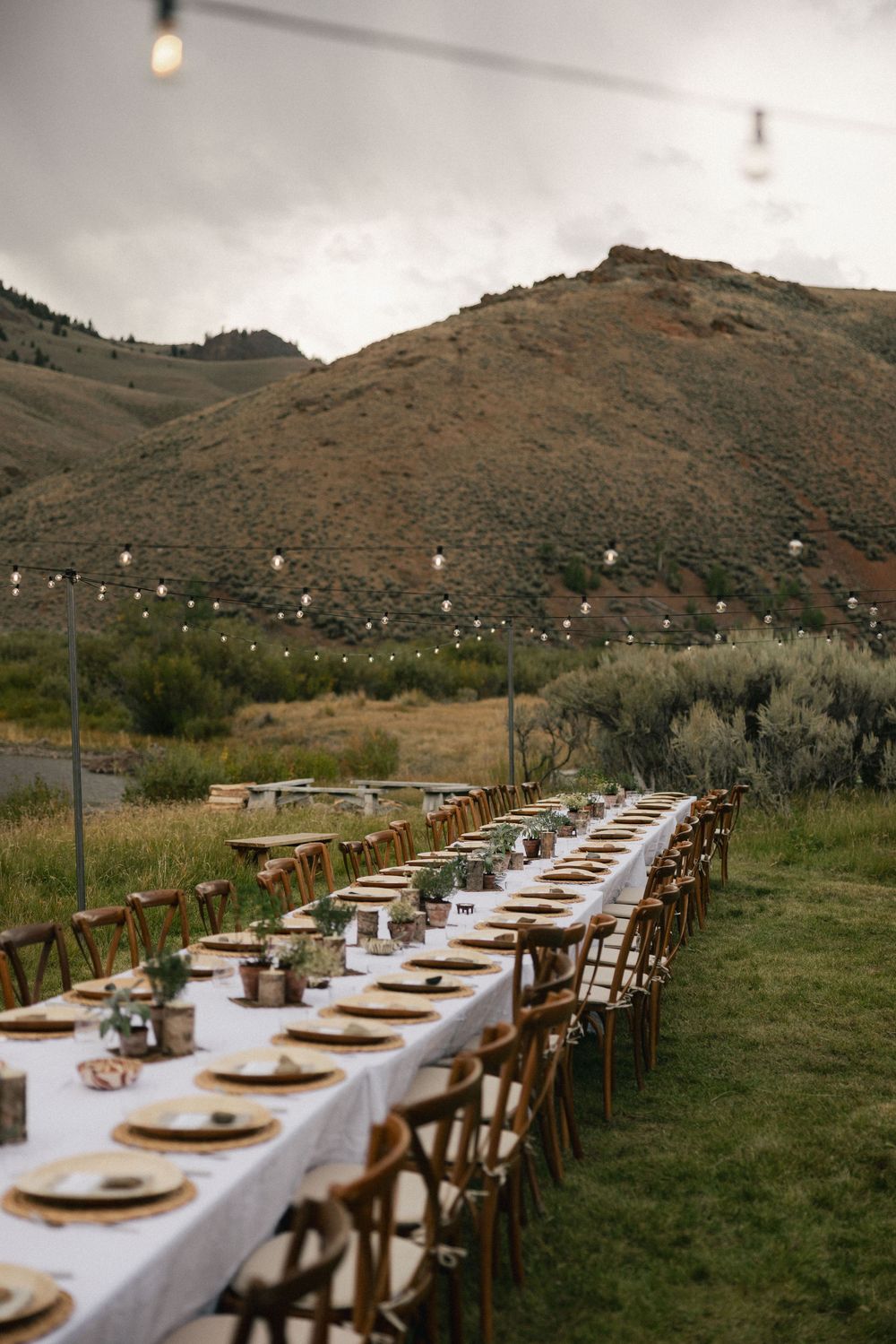 Long outdoor dining table set for a wedding reception with string lights and scenic mountain backdrop.