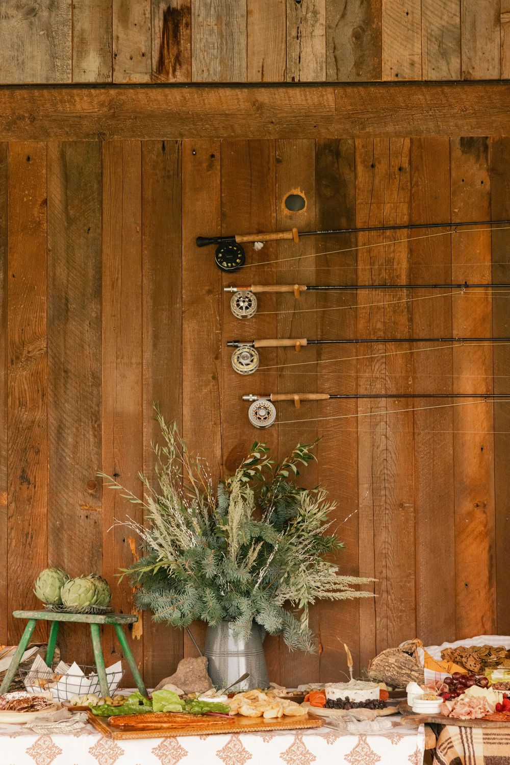 Rustic barn wall display with fishing rods, floral arrangement, and charcuterie spread on a patterned tablecloth.