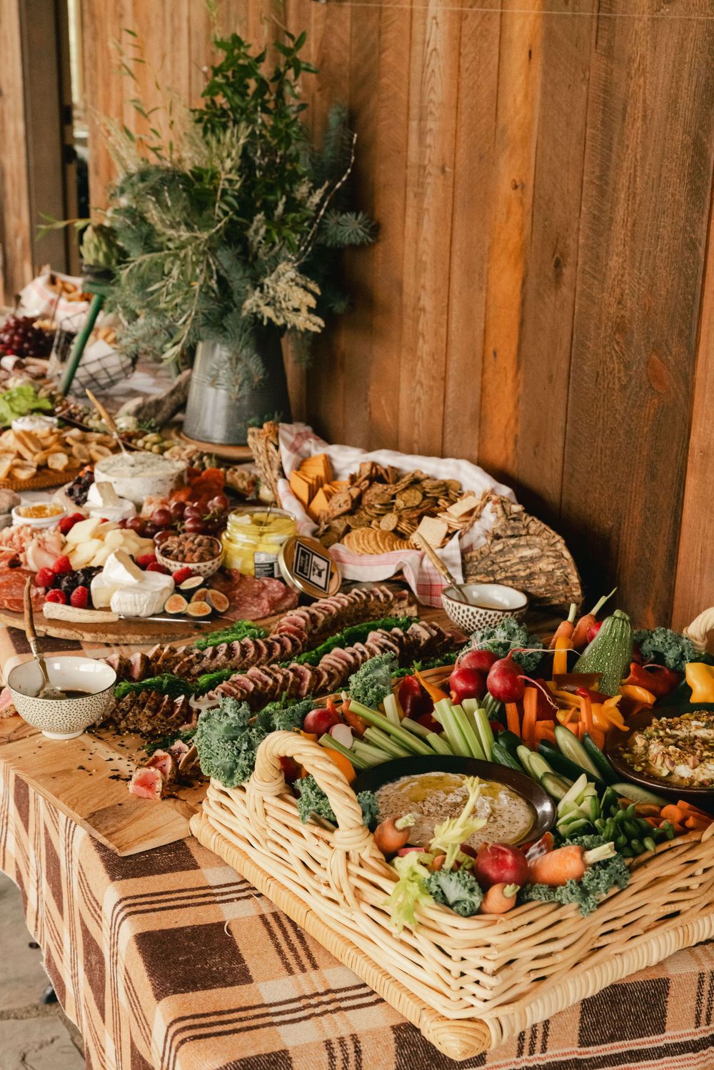 Rustic grazing table with charcuterie, fresh vegetables, cheeses, and breads displayed in wicker baskets on a plaid tablecloth.