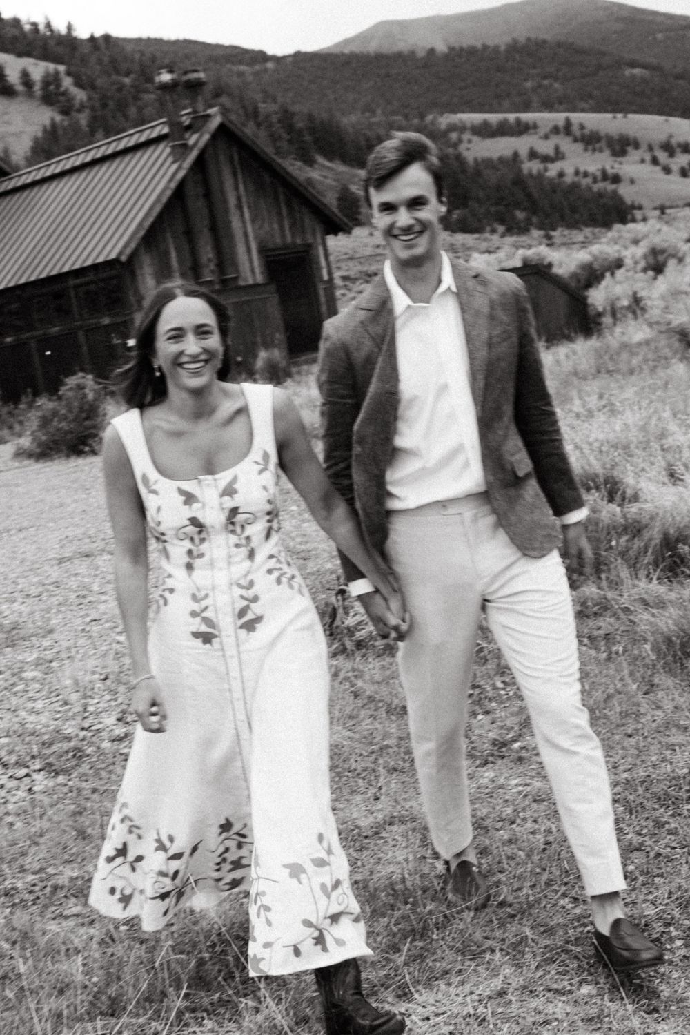 Happy couple holding hands walking outdoors near rustic barn, woman in floral dress, man in blazer, black and white photo.