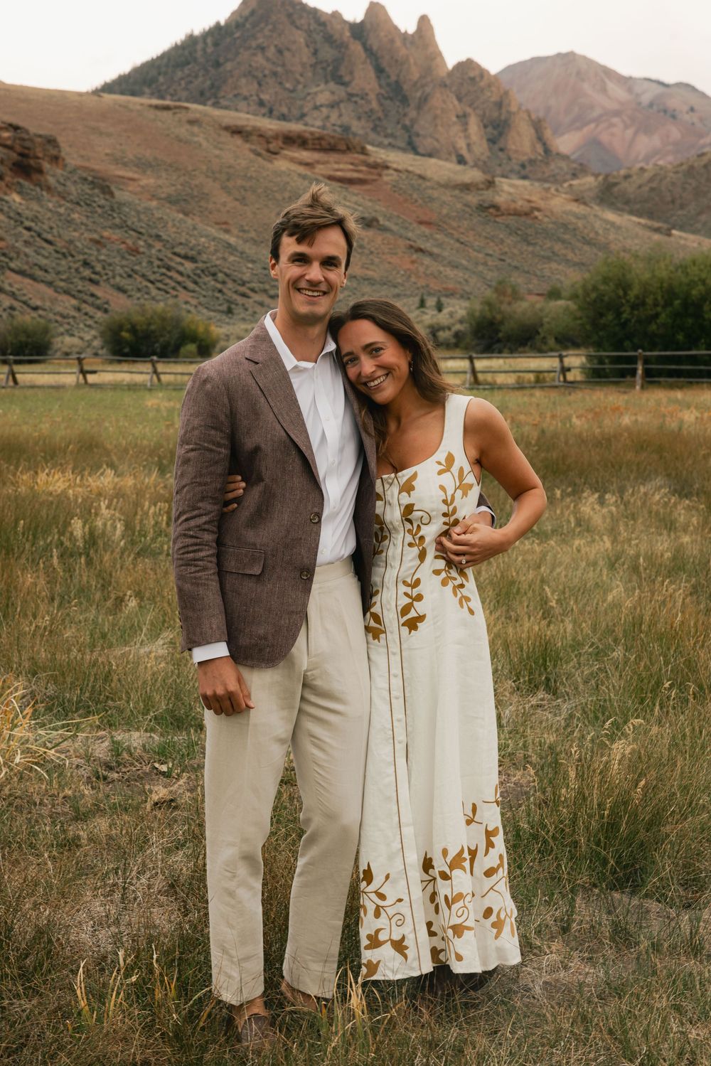 Couple posing in a field with rocky mountain backdrop, man in brown blazer and woman in floral white dress.