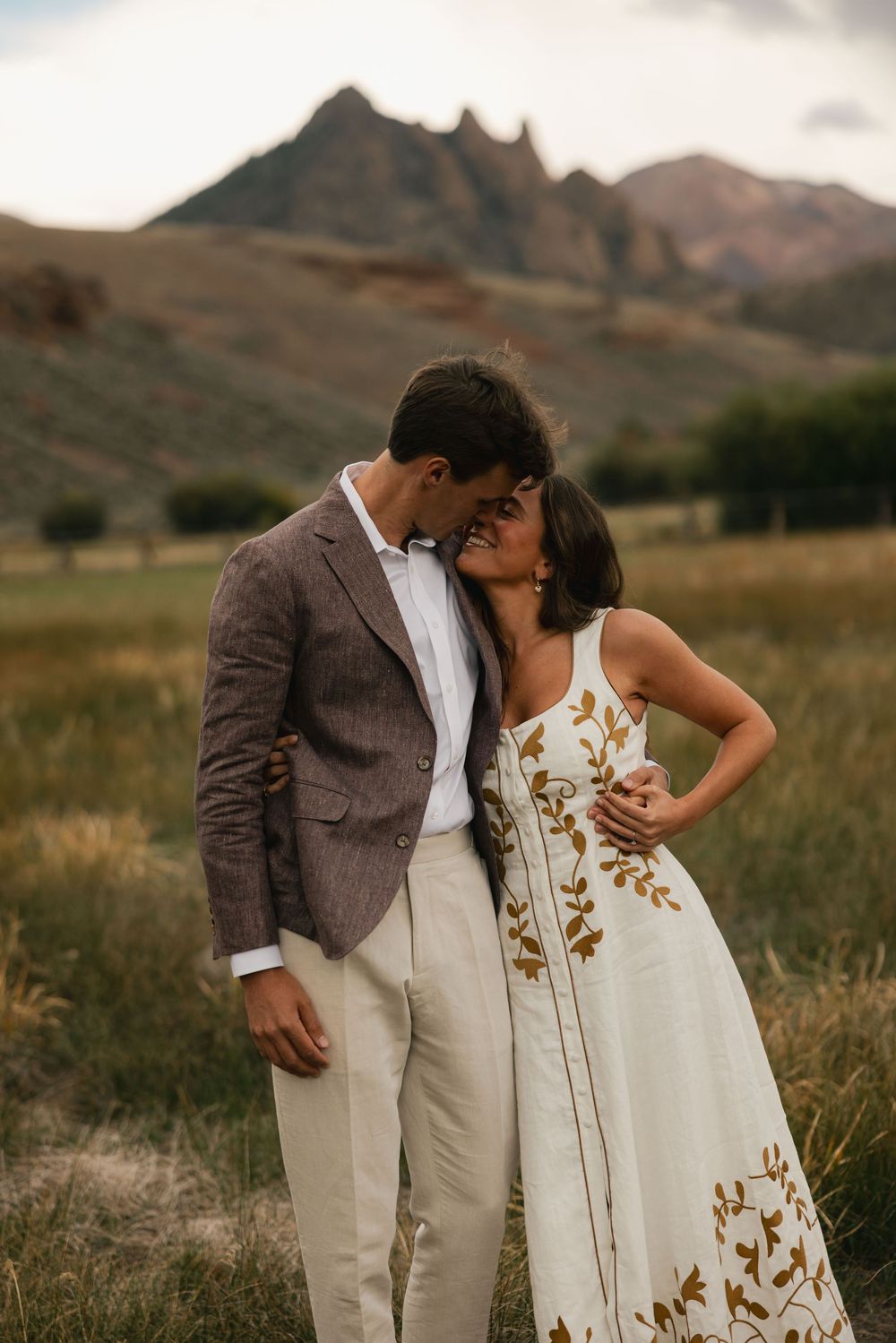 Couple sharing a romantic moment in a field with mountain backdrop, man in brown blazer, woman in white floral dress.