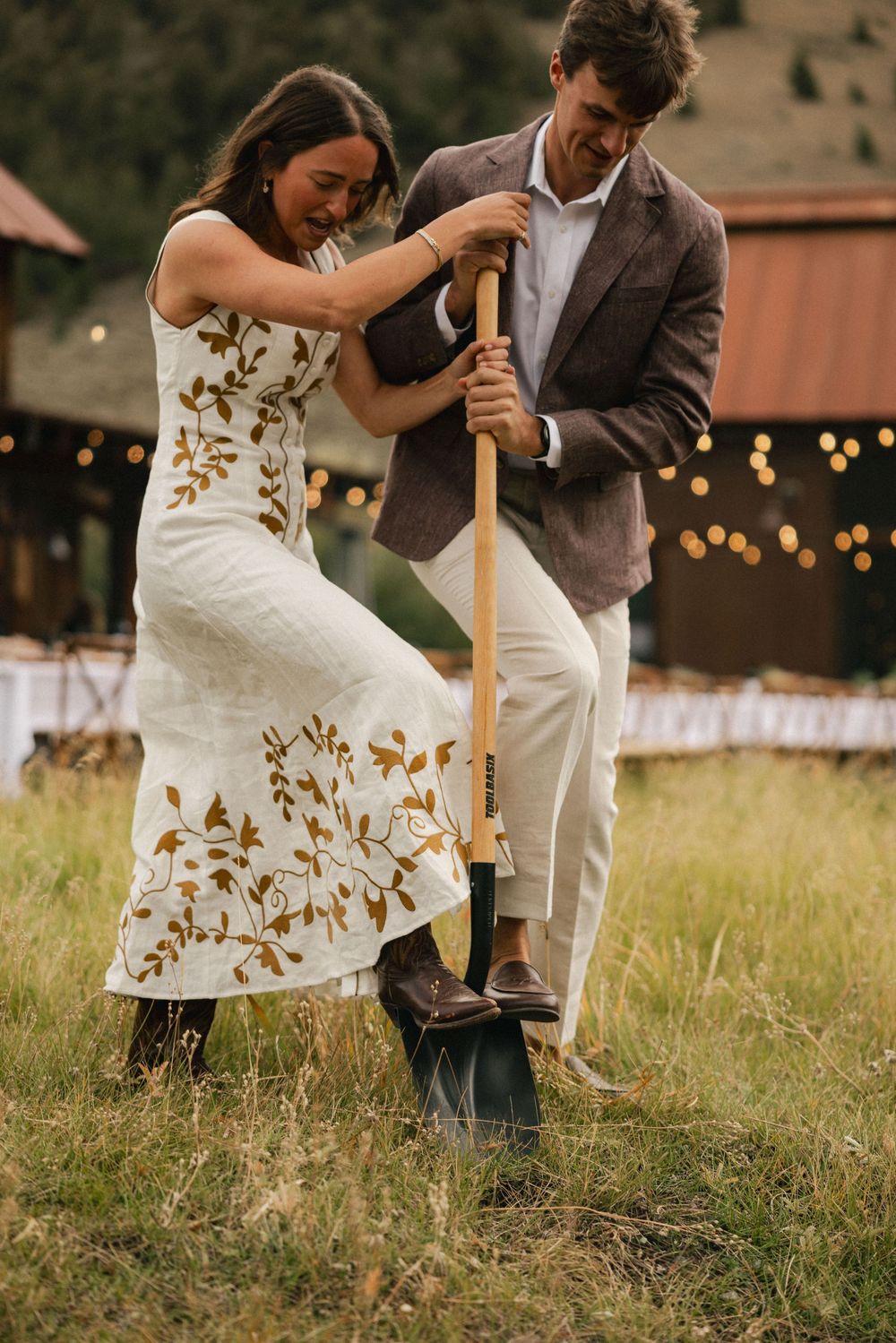 Couple in wedding attire digging with a shovel in a grassy field with string lights and barn in background.