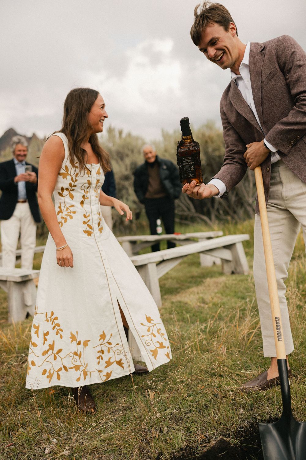 Couple laughing while burying a whiskey bottle outdoors, a wedding tradition, with guests in the background.