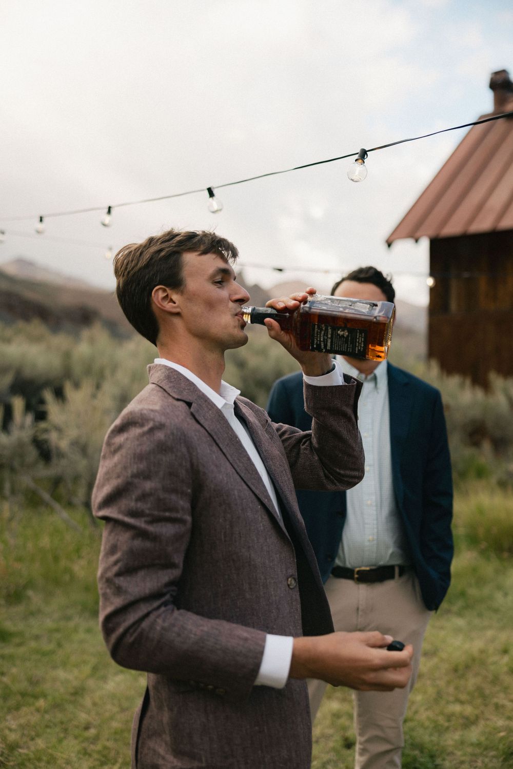 Man in brown suit drinking from whiskey bottle outdoors at rustic event with string lights and mountains.