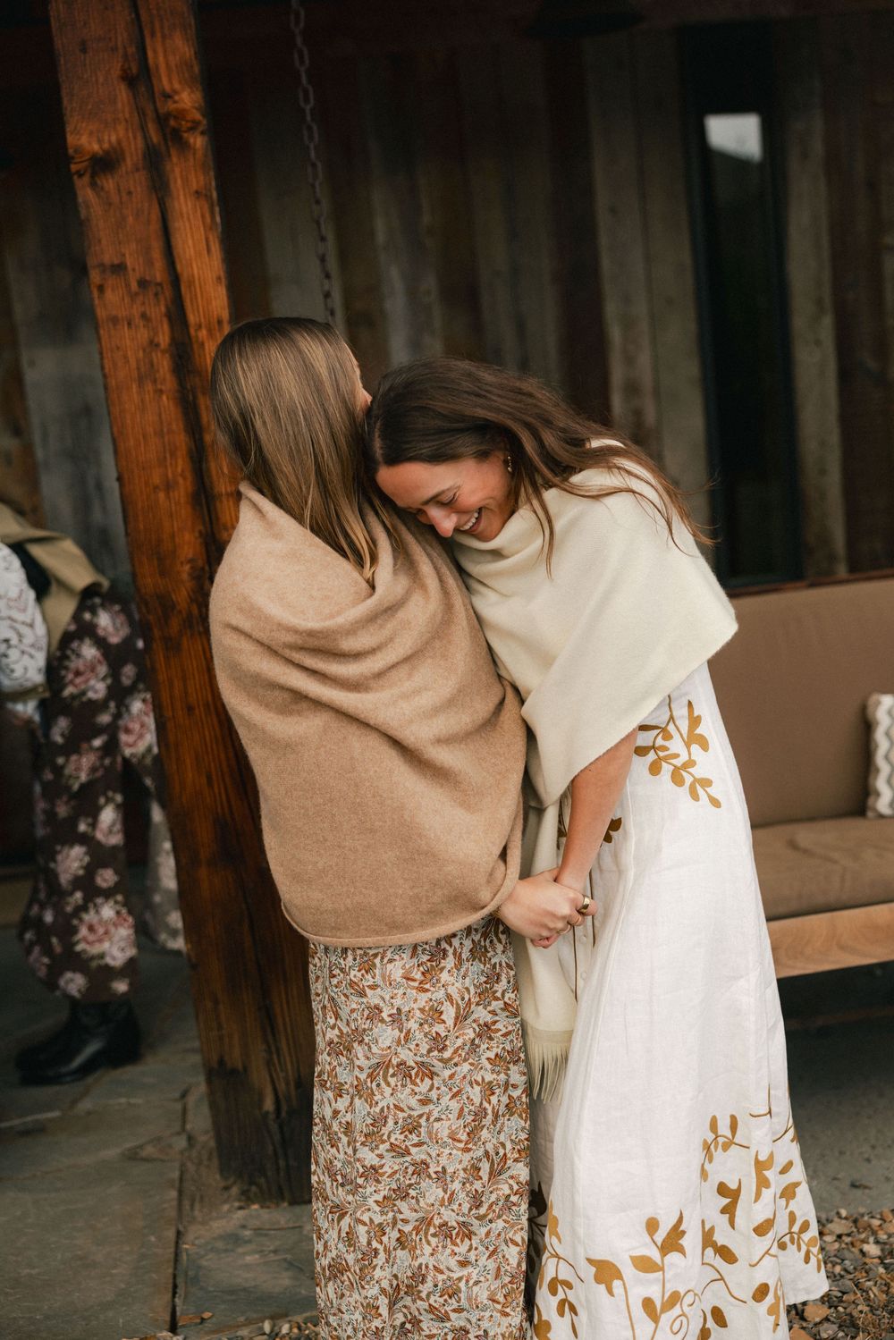 Two women wrapped in cozy shawls embrace warmly outdoors near a rustic wooden structure.