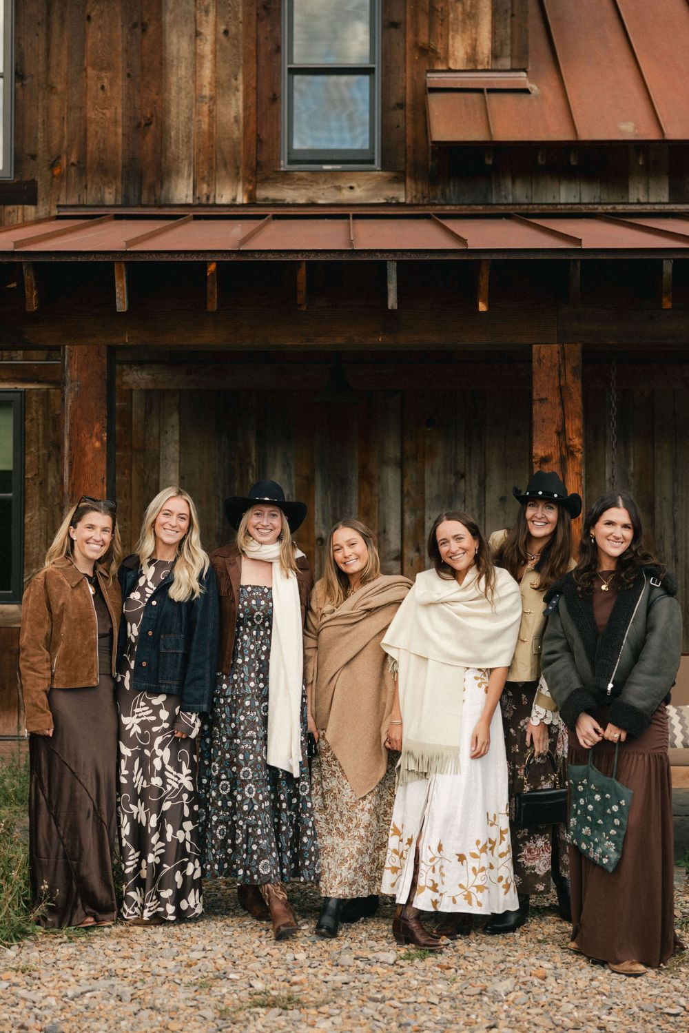 Seven women in bohemian fall outfits pose together outside a rustic wooden barn with a metal roof.