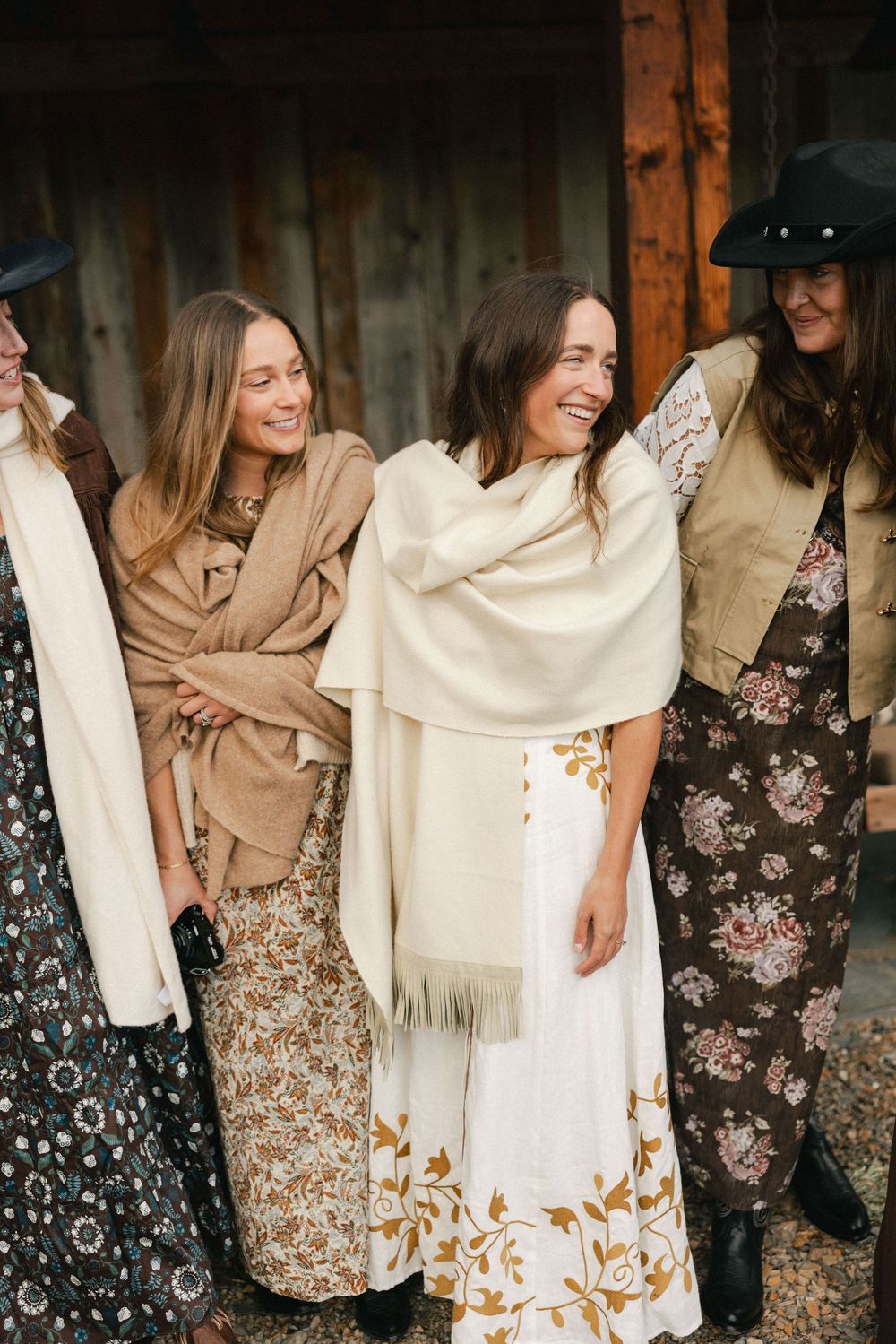 Group of women in boho floral maxi skirts and neutral wraps smiling together near a rustic barn.
