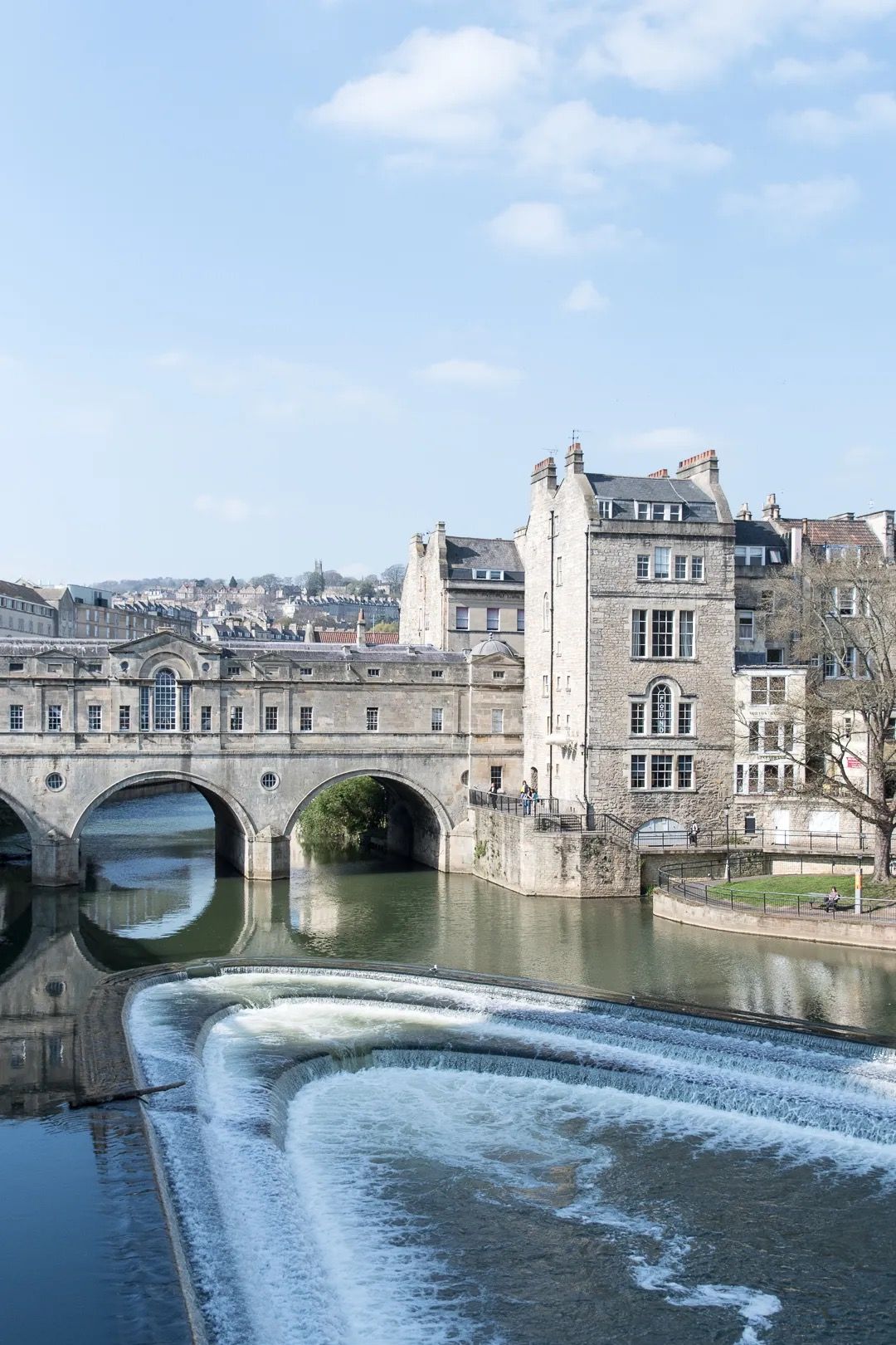 Historic stone bridge over a river with a curved weir and city buildings in the background.