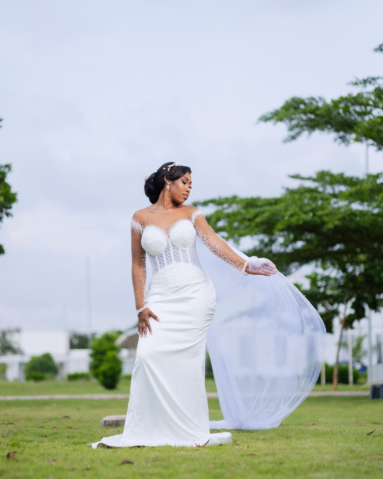 A bride in a flowing white wedding dress poses gracefully on a grassy hill with trees in the background.