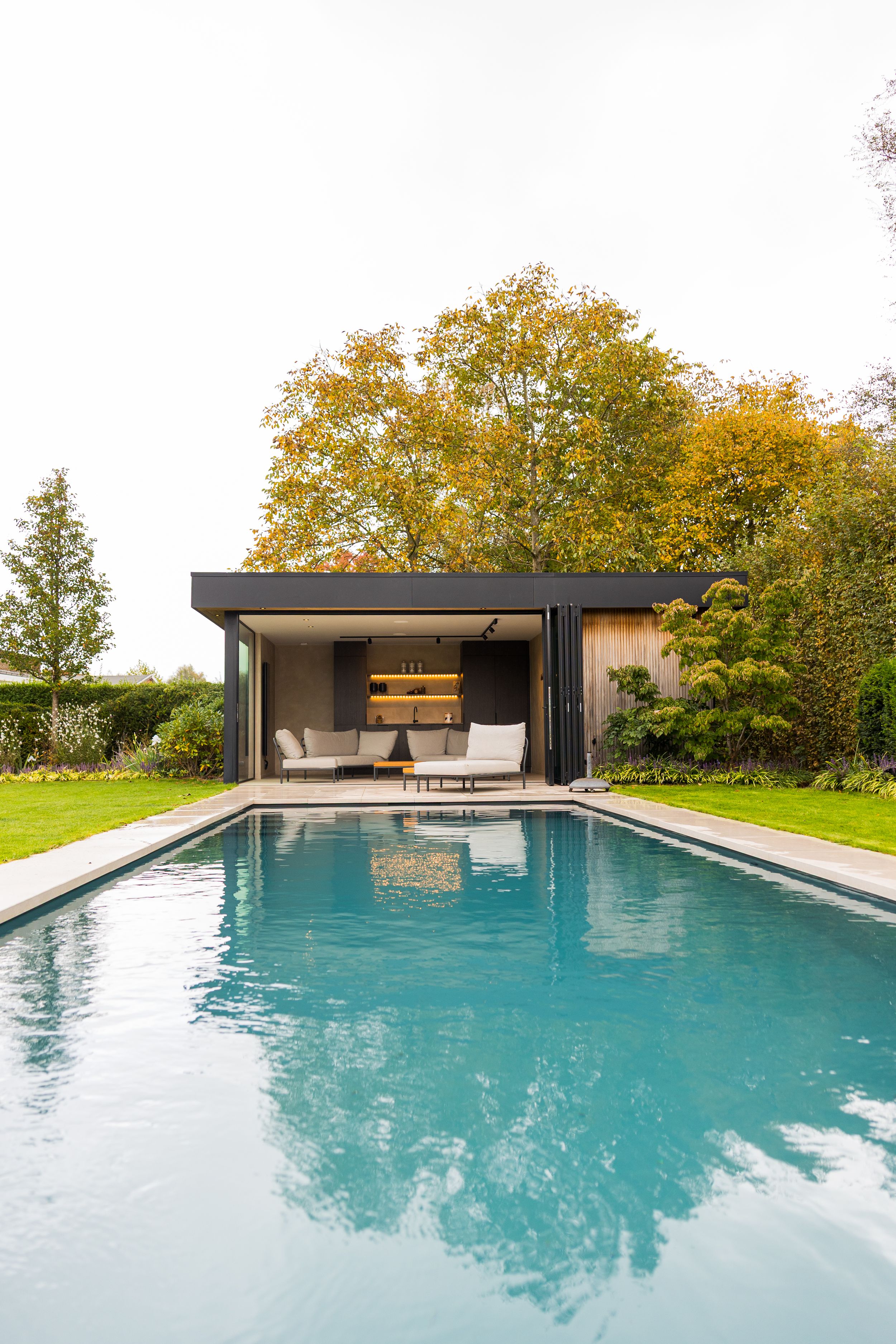 Modern pool house with sleek design featuring turquoise water and autumn trees in the background.