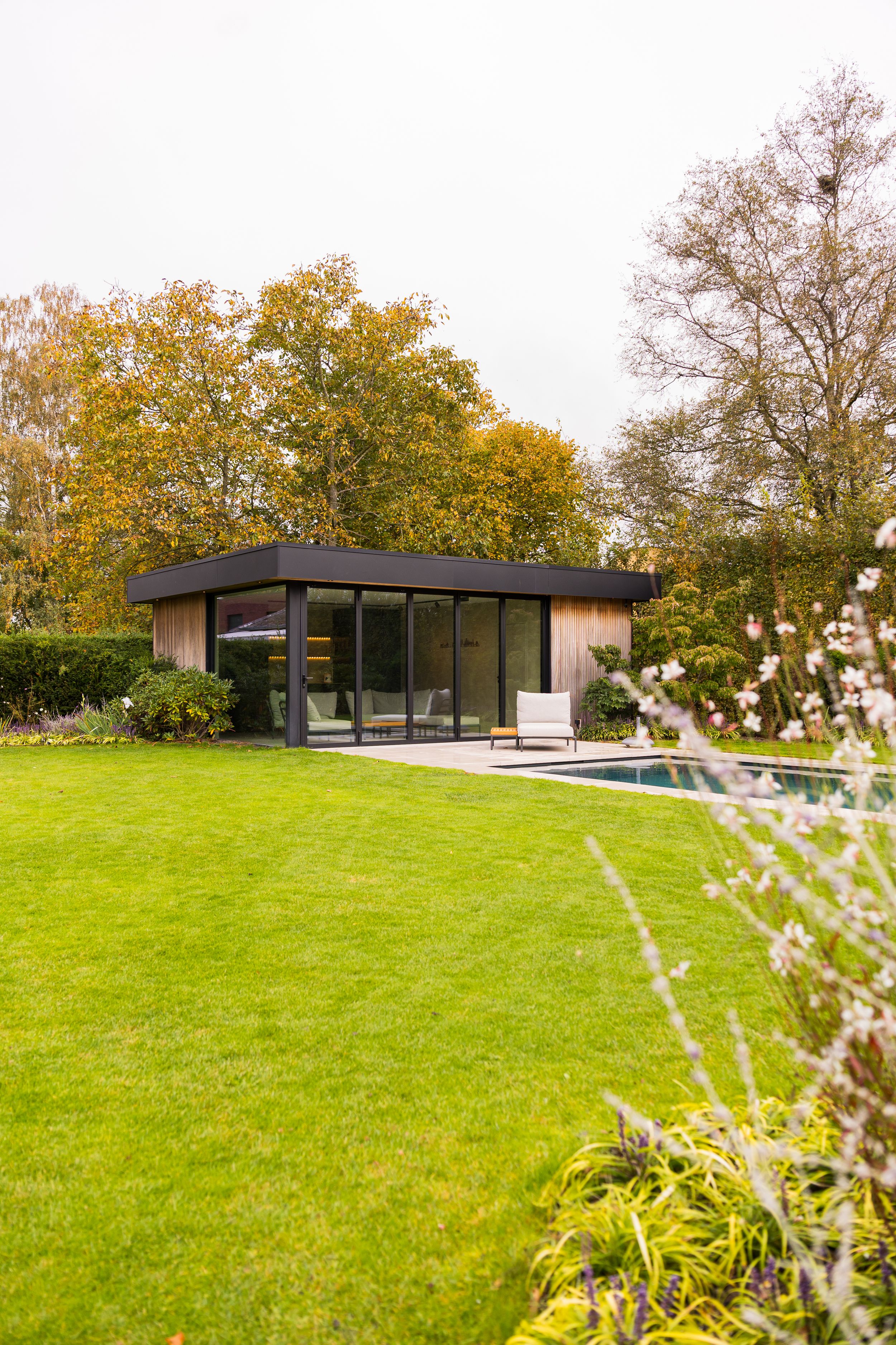 Modern minimalist pool house set on a sprawling green lawn with autumn trees.