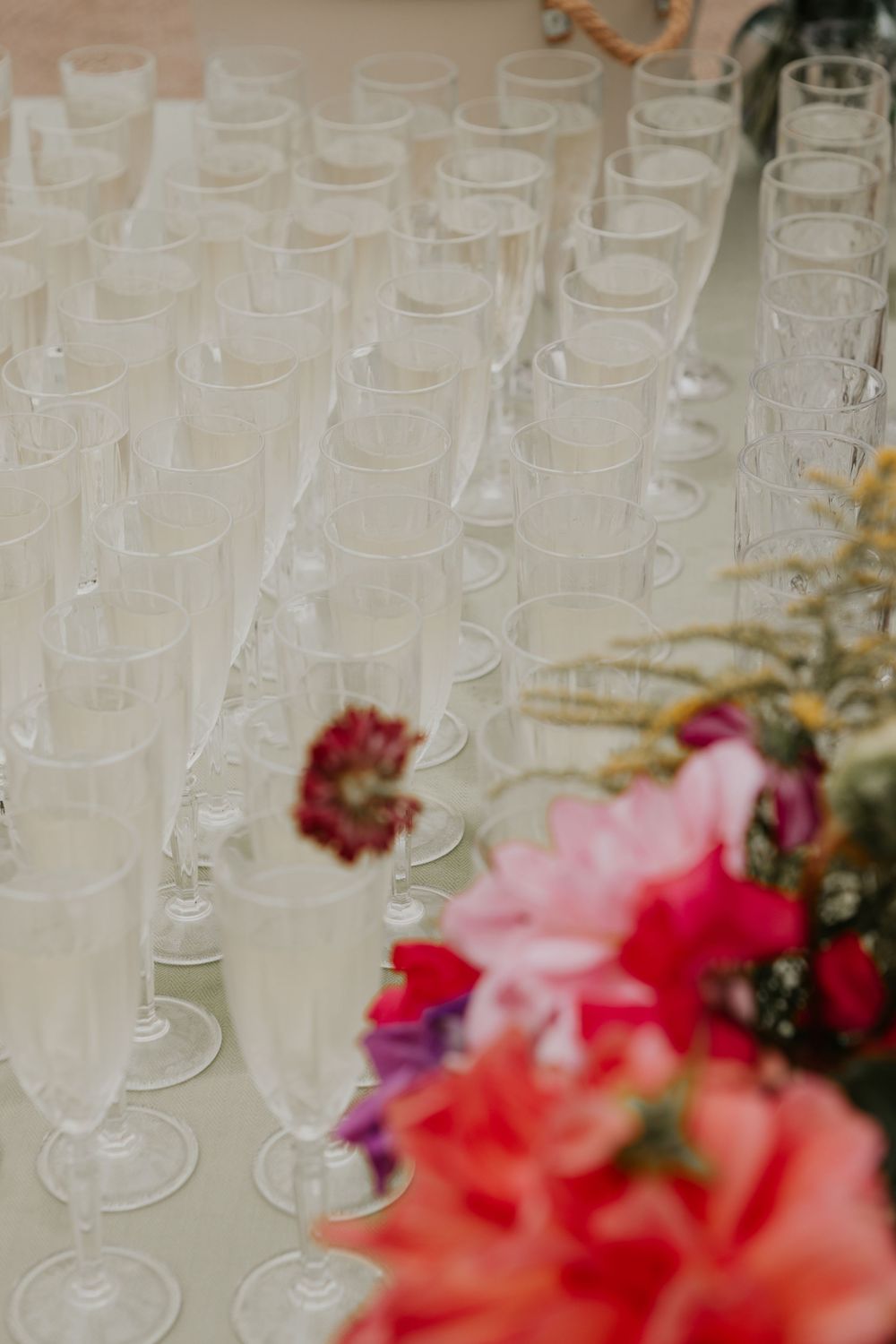 Rows of empty champagne flutes arranged on table with colorful pink and red floral arrangement.