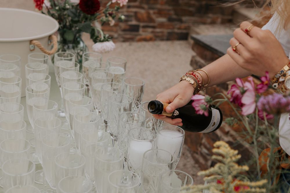 Wedding staff member pouring champagne into rows of glasses at outdoor wedding reception drinks table.