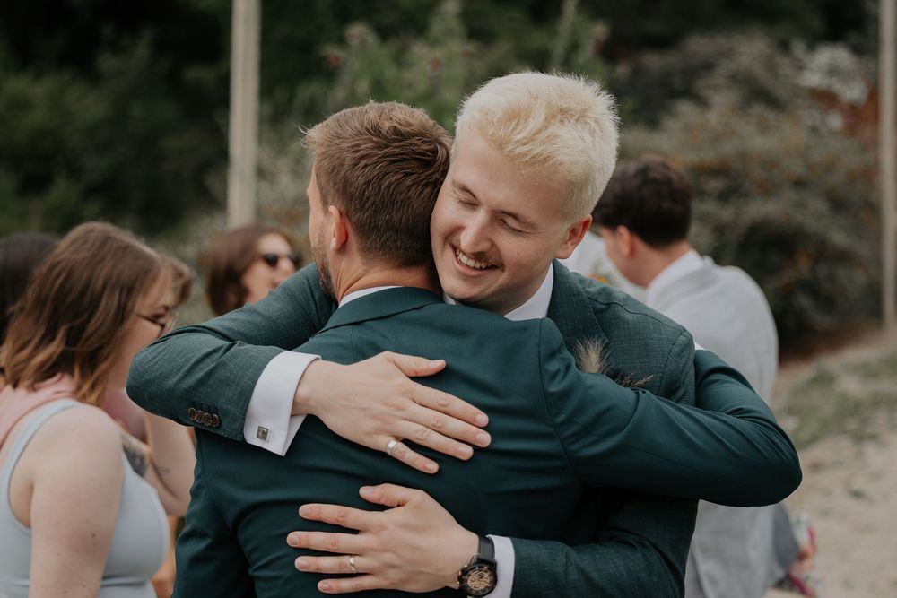 Two men in green suits share a warm embrace at an outdoor wedding celebration.