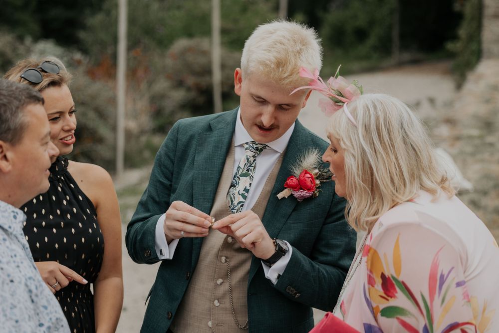 Blond groom in green suit examines boutonniere with smiling wedding guests outdoors.