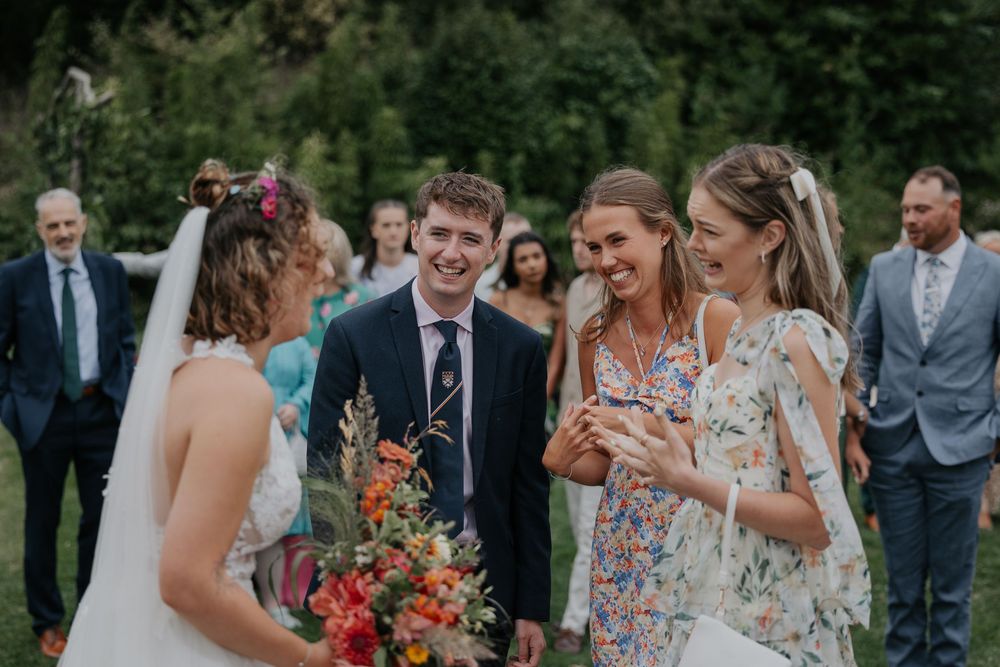 Bride with colorful bouquet laughs with guests in floral dresses at outdoor wedding.