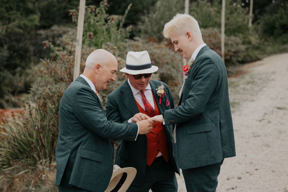 Three men in formal suits examine wedding rings outdoors on a sunny day.