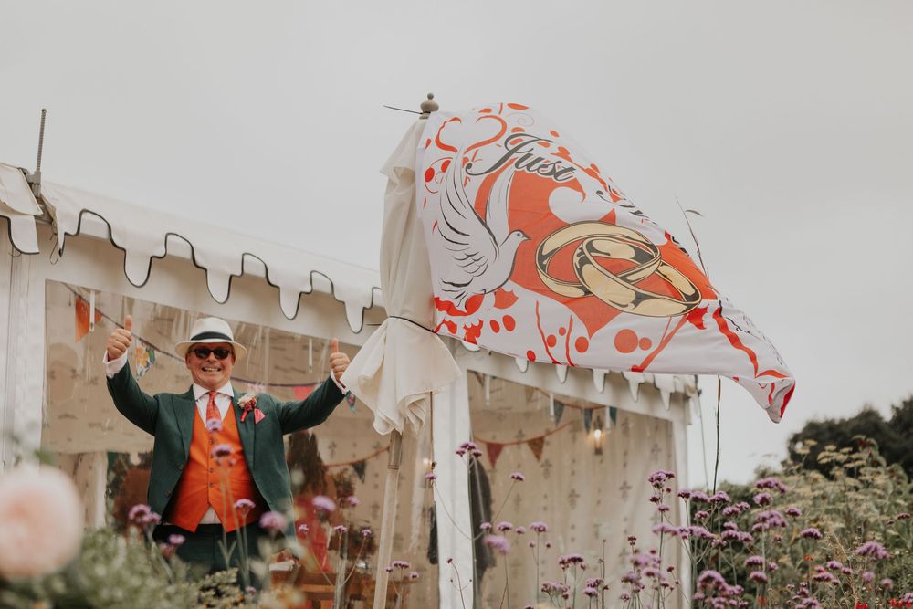 Festive man in orange vest and hat waves colorful wedding flag outside decorated tent.