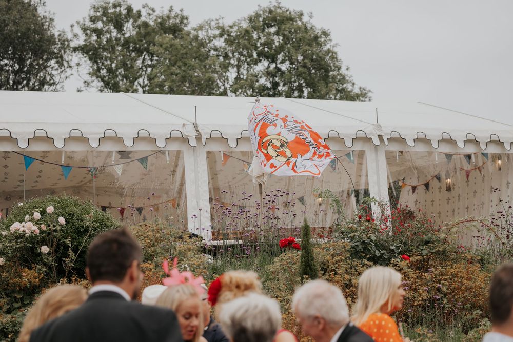 Wedding guests gather outside white tent adorned with colorful flag and bunting decorations.
