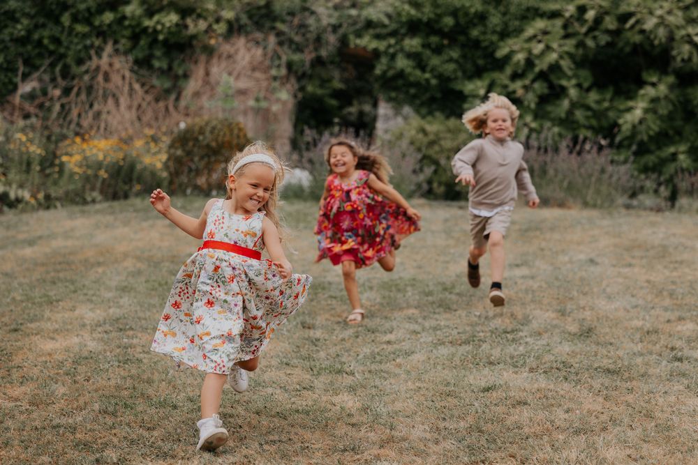 Three young children in colorful dresses run and play joyfully on a green lawn.
