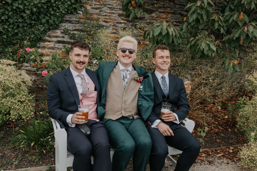Three young men in suits smile while holding drinks on a bench at a wedding.