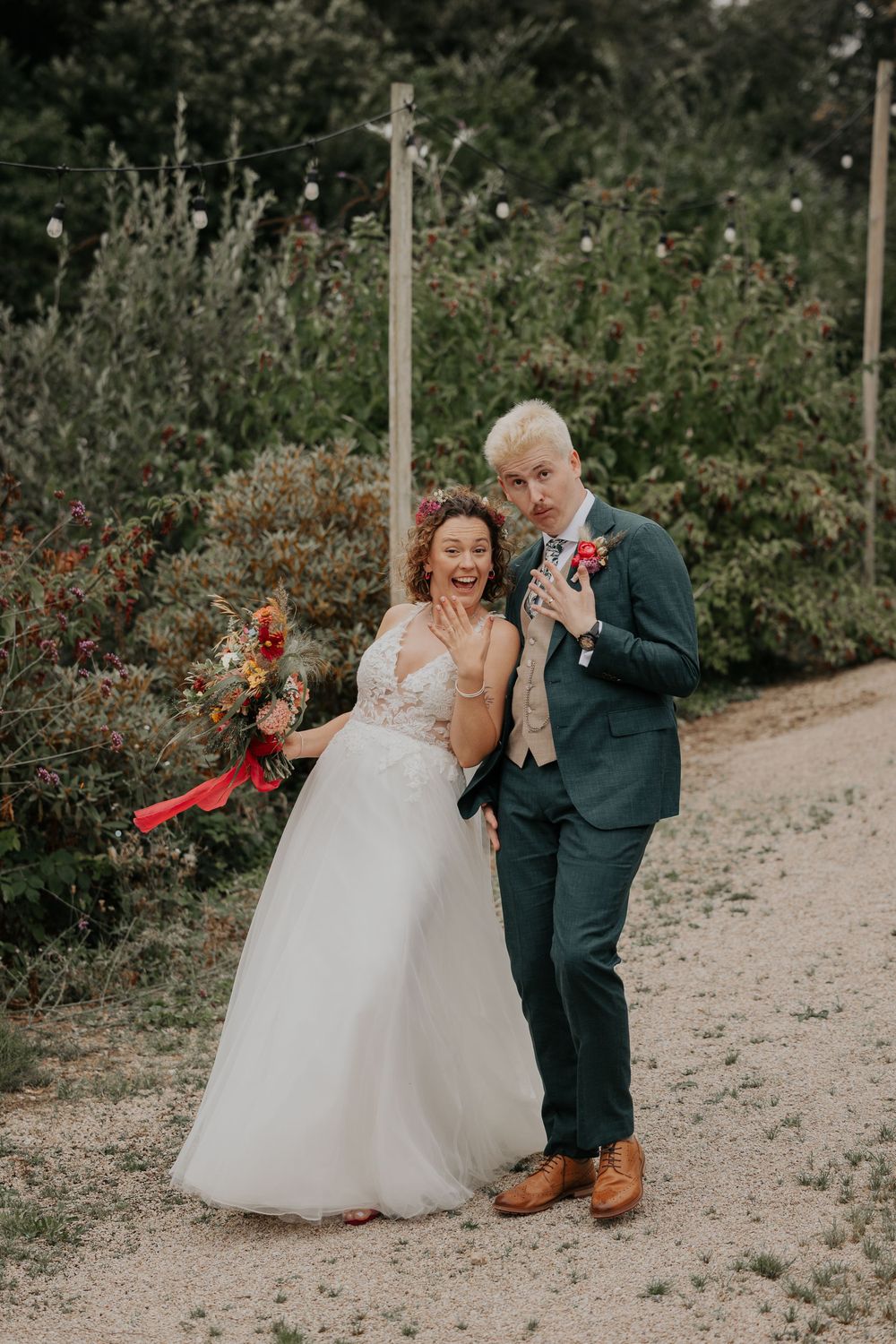 Joyful bride and groom laughing together on a garden path, bride holding colorful bouquet with red ribbons.