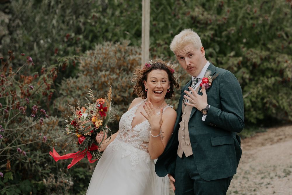 Happy bride laughing with groom in teal suit outdoors, bride holding vibrant autumn wedding bouquet.