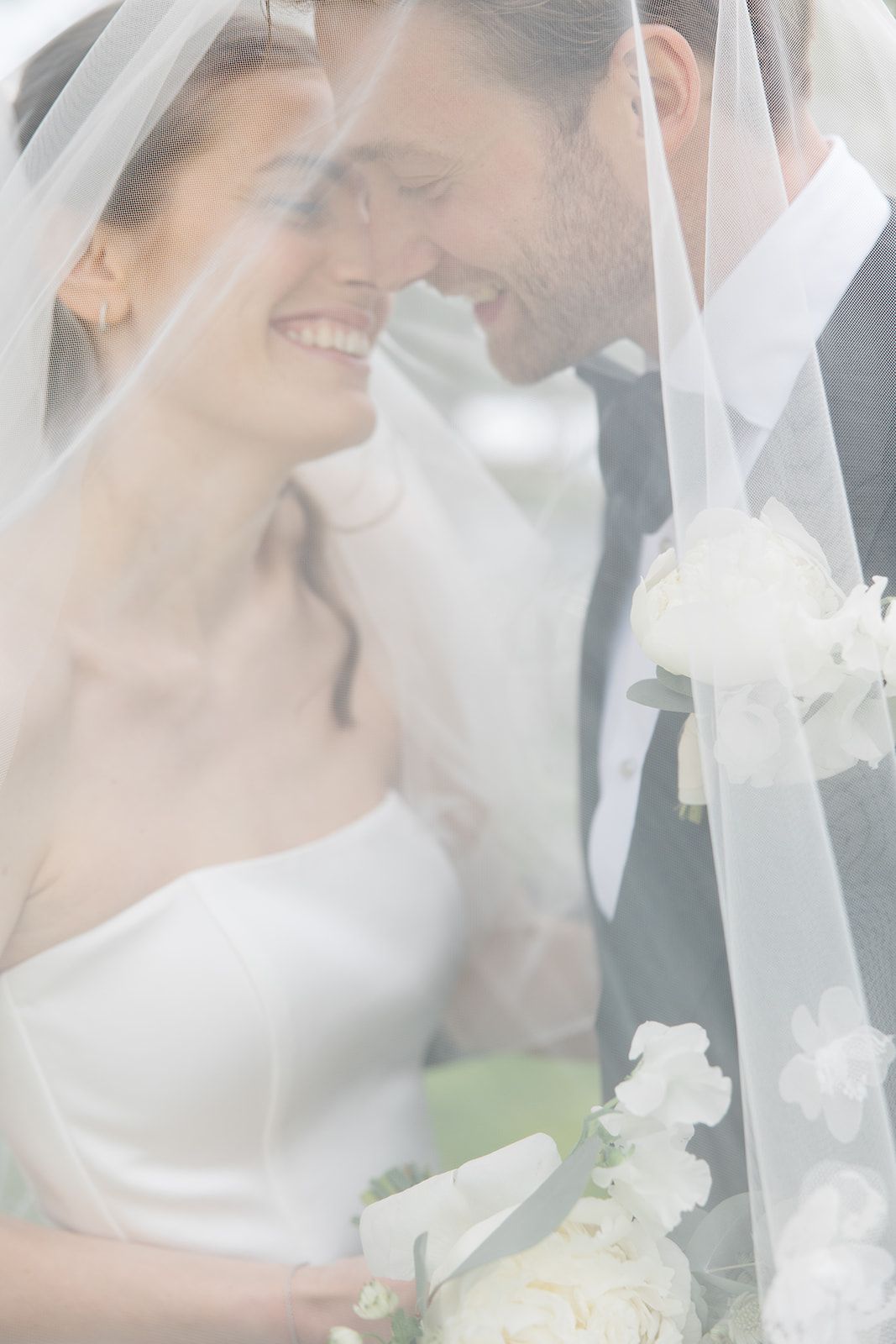 Romantic wedding couple shares intimate moments under a flowing white veil while holding a bouquet of white flowers.