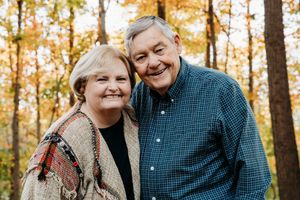 A couple embraces while sitting outdoors among autumn trees with golden foliage in the background.