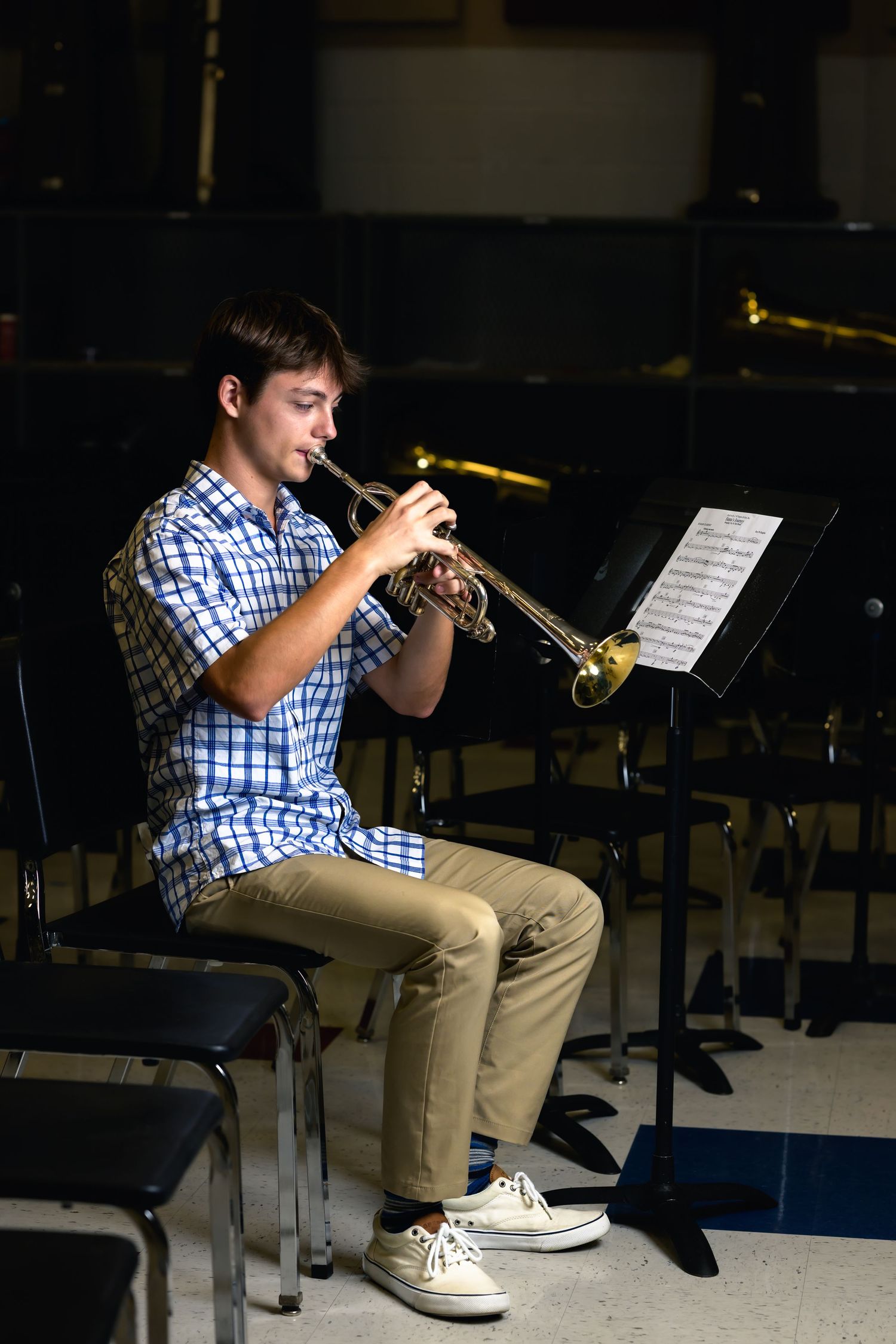 A musician in a checkered shirt plays trumpet while seated in a practice room with music stands visible.