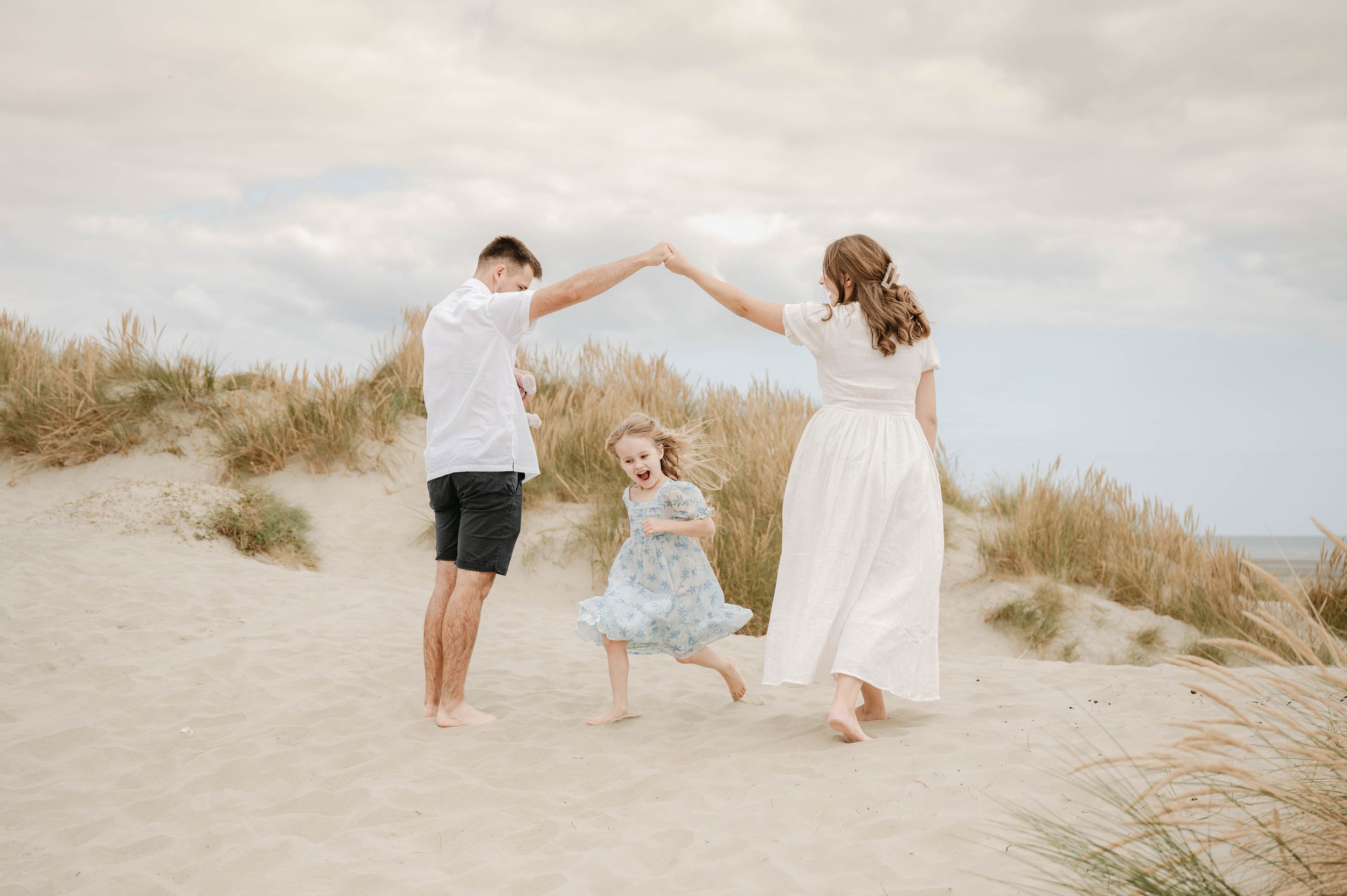 A romantic couple in white outfits dances playfully together on a sandy beach dune at sunset.