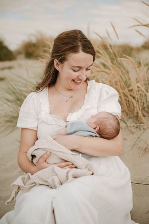 Mother in white dress holding sleeping newborn baby on sandy beach with tall grass in soft natural light.